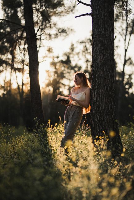 Young woman reading a book while leaning against a tree in a sunlit forest.