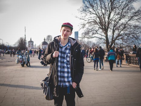 白天，一个穿着格子衬衫、戴着帽子的年轻人在繁华的城市公园里悠闲地散步