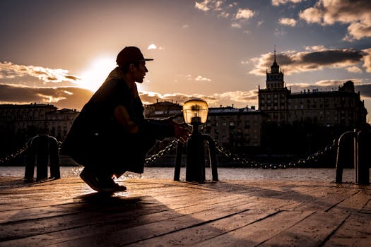 日落时分，一名戴帽子的男子蹲在城市河边的码头上，构成了一幅富有戏剧性的城市景象