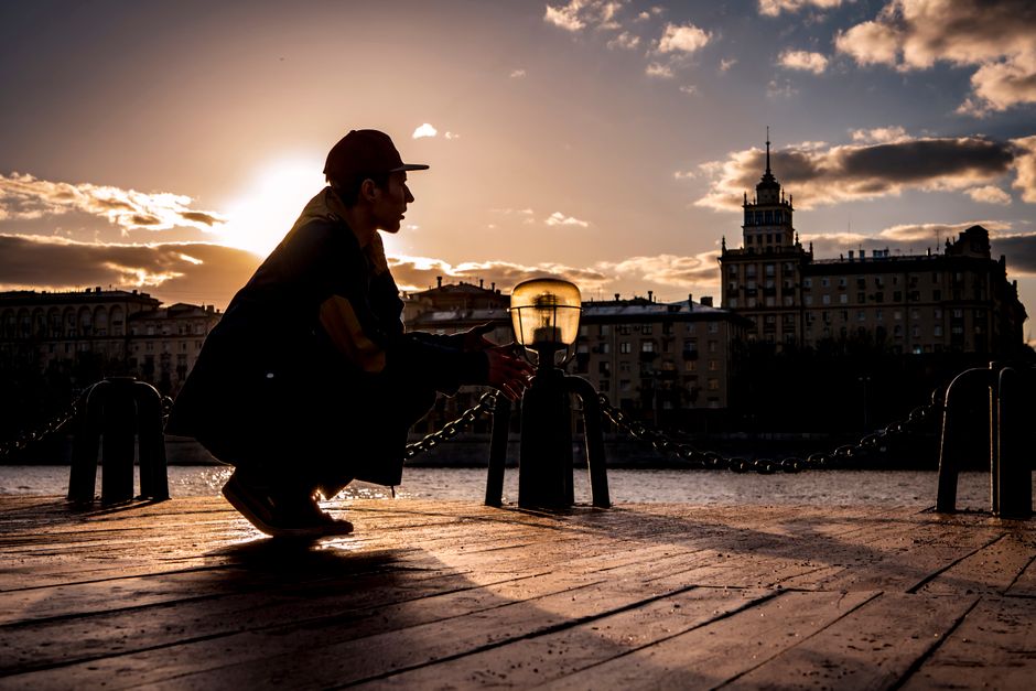 Silhouette of man in cap squatting on a pier by city river during sunset, creating a dramatic urban scene.