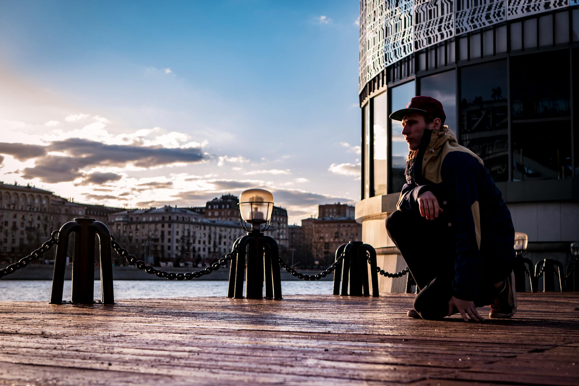 A person kneels on a pier by a city river at sunset, with modern buildings in the background.
