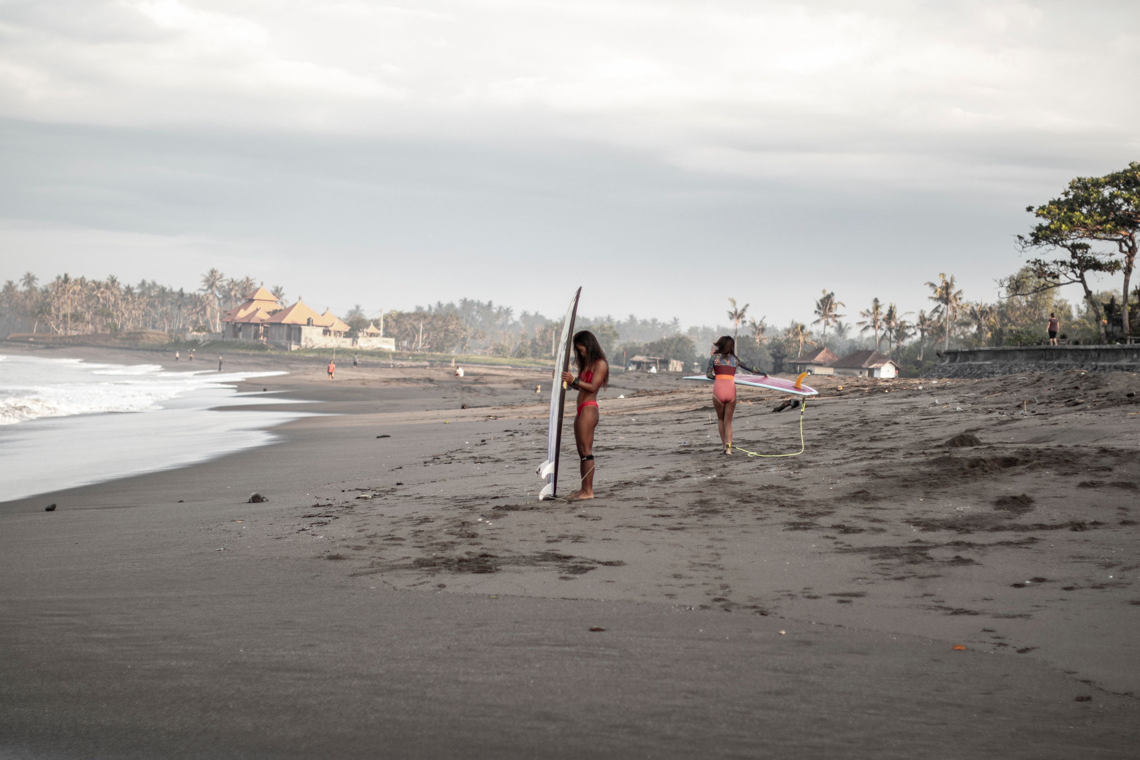Two women with surfboards on a tropical beach at sunrise, preparing for surfing adventure.