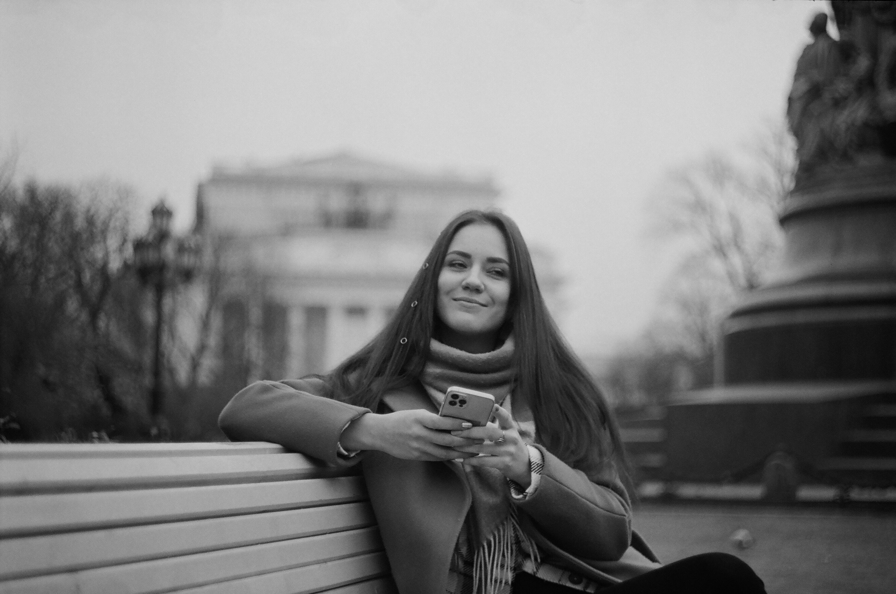 A smiling woman with long hair sits on a bench in a park, using her smartphone.