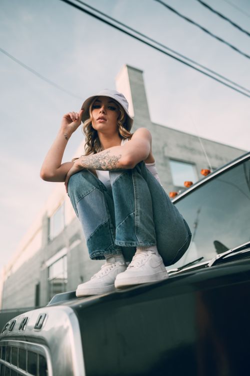 Free Stylish woman in a bucket hat and jeans sitting on a truck hood in an urban setting. Stock Photo
