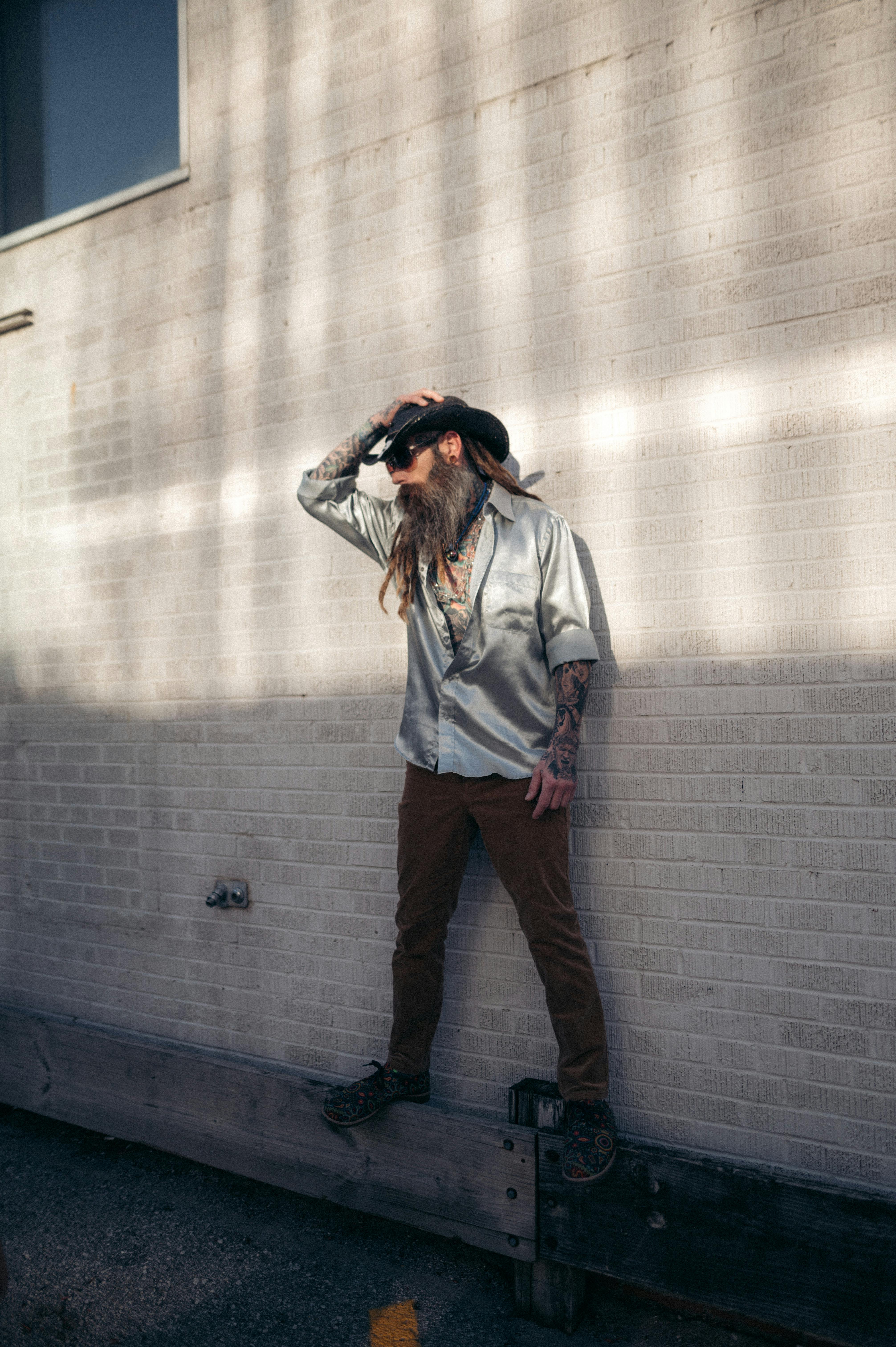 Bearded man in casual attire stands confidently against a sunlit brick wall.
