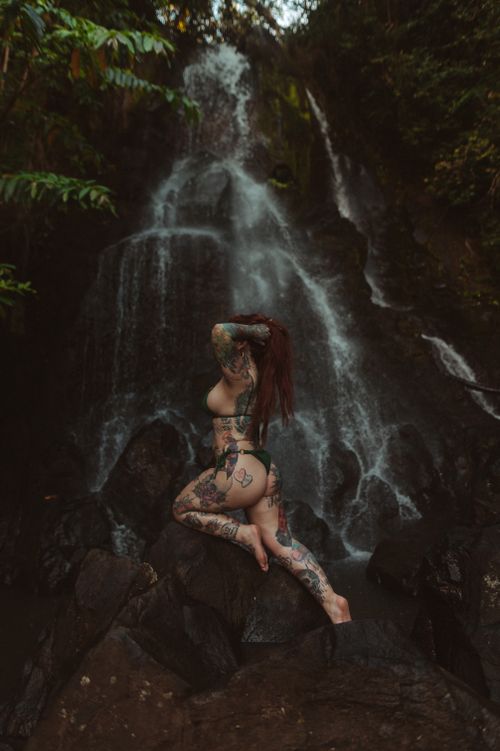 Free A tattooed woman in swimwear poses gracefully by a waterfall amidst a natural rock formation. Stock Photo