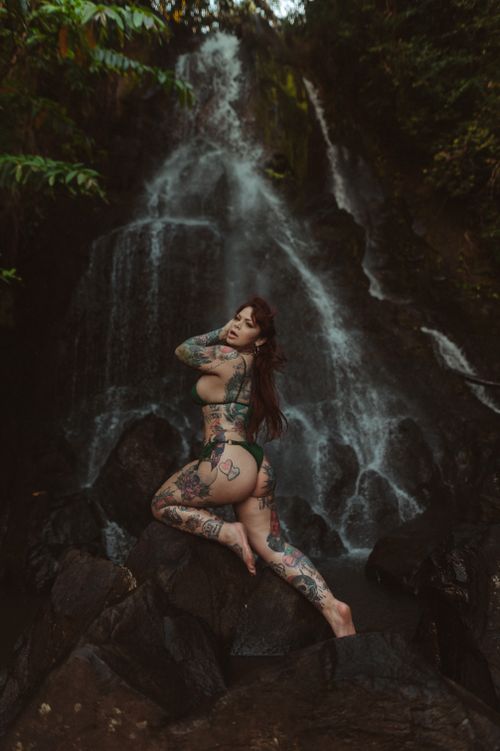 Free A tattooed woman in a bikini poses confidently by a lush waterfall setting. Stock Photo