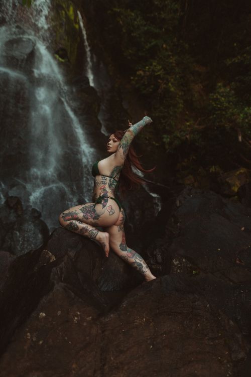 Free A tattooed woman in swimwear posing dramatically on rocks by a waterfall. Stock Photo