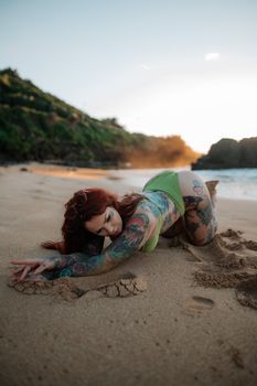 A tattooed woman in bikini poses on a sandy beach during the day.