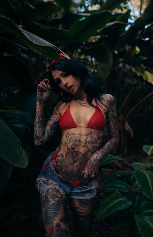 Free Tattooed woman in a red bikini with denim shorts posing outdoors in lush greenery. Stock Photo