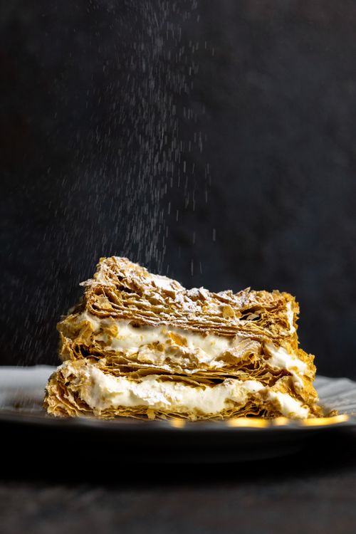 Free A close-up of layered mille-feuille pastry sprinkled with powdered sugar, served on a plate. Stock Photo