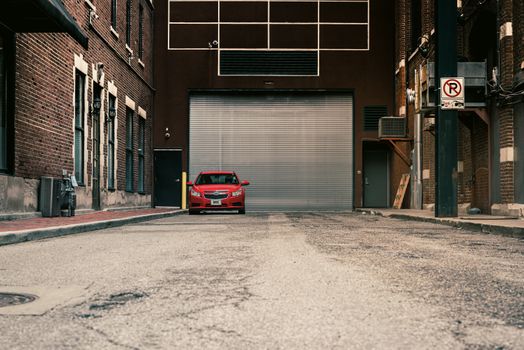 A red car parked in an urban alley surrounded by brick buildings and a garage door.