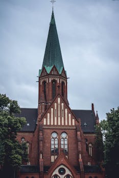 一座哥特式红砖大教堂高耸入云，尖顶直指多云的天空，四周绿植环绕