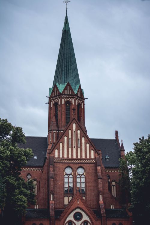 Free A red brick Gothic cathedral with a towering spire under a cloudy sky, surrounded by greenery. Stock Photo