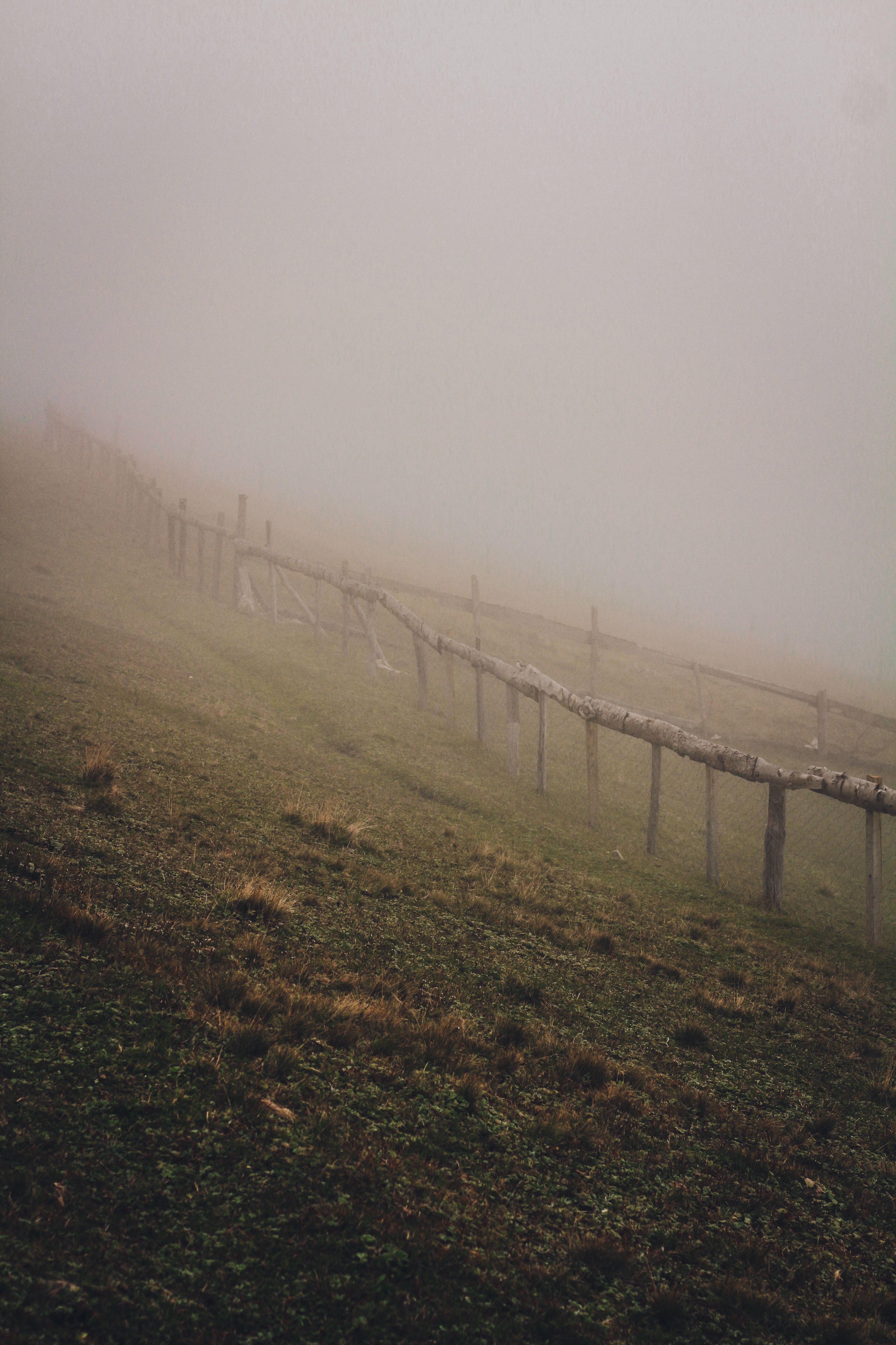 A foggy scene of a sloping field with a wooden fence and green grass.