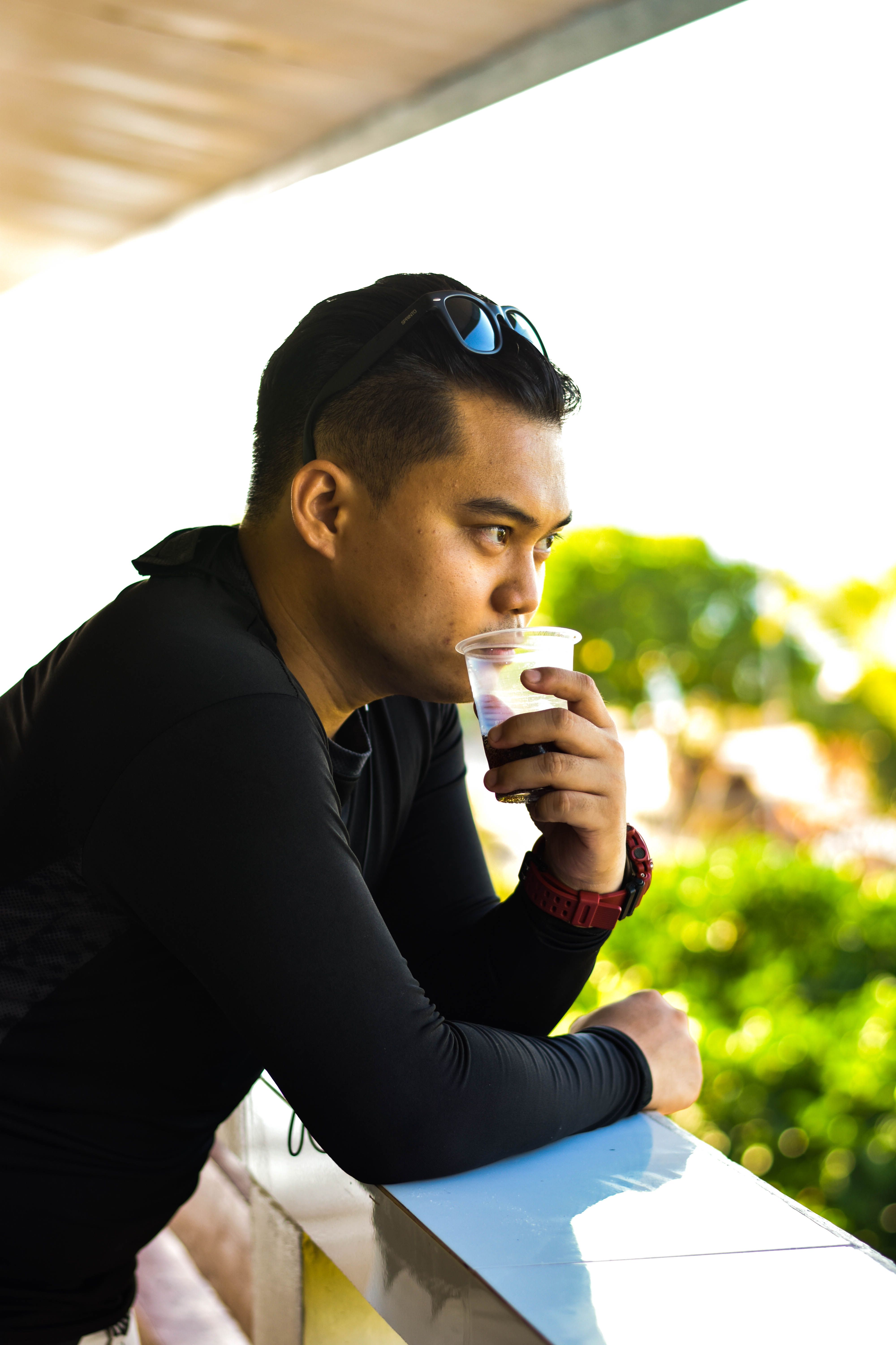 Man in black long sleeves leans on a balcony, enjoying a drink from a plastic cup.