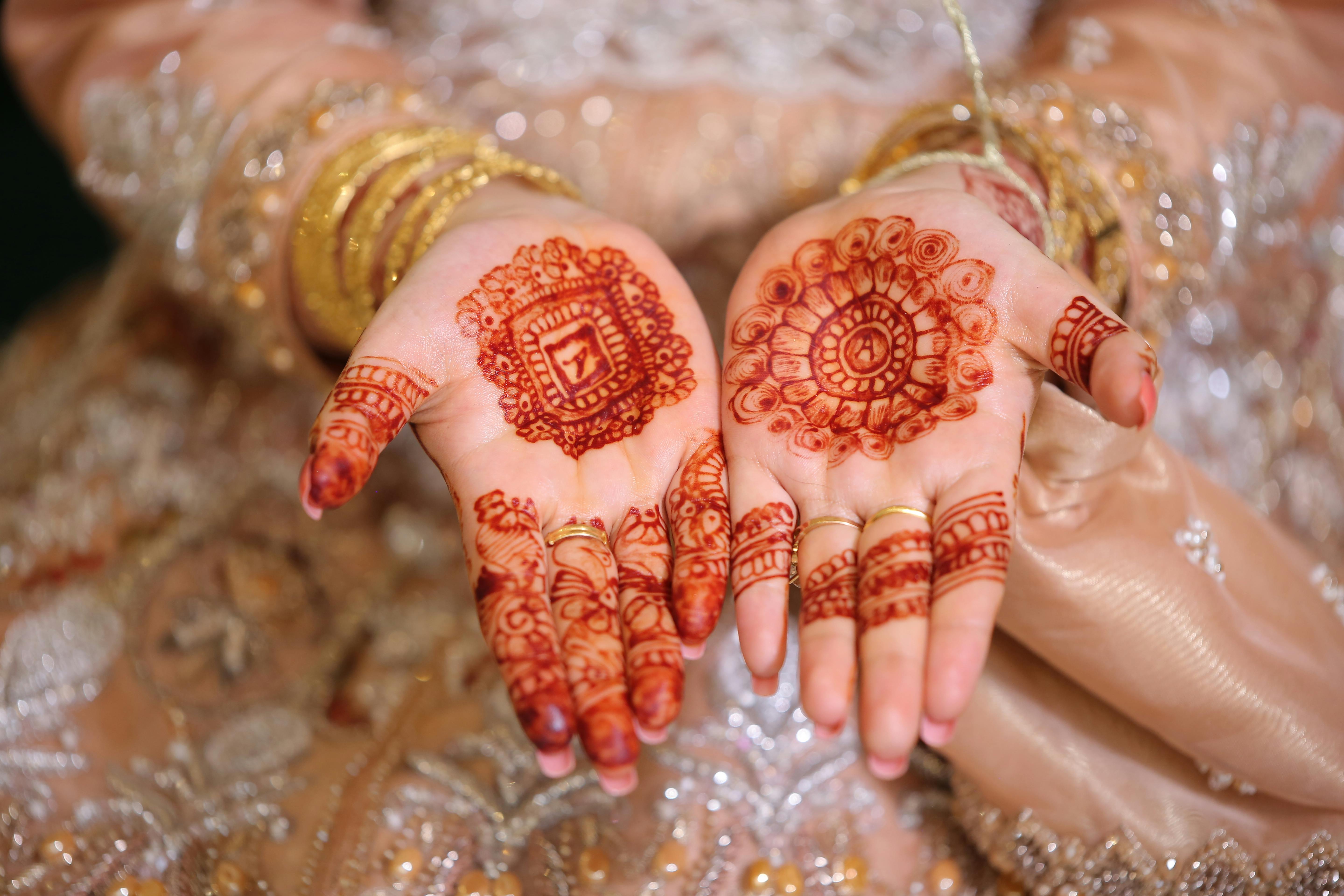 The close-up shot of Indian bride with beautiful red saree showing mehndi ( henna) tattoo hand with a lot of glitter bracelets (bangles) on her wrist  and wedding rings by Manee S. Photo