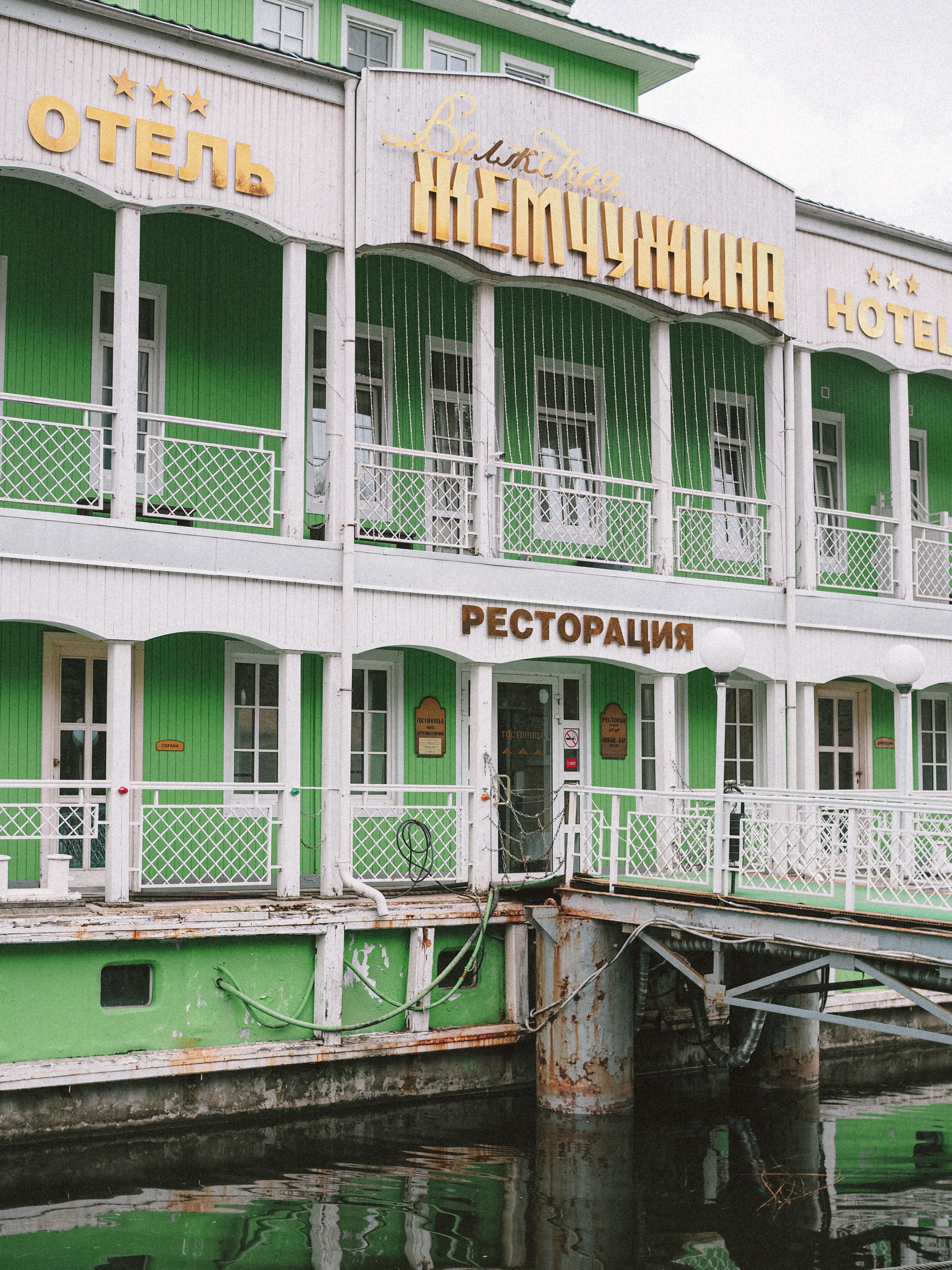 Photograph of a green riverside hotel with Cyrillic signs reflecting in water.