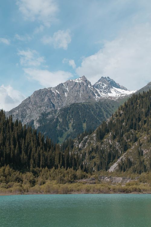 Free Majestic mountain landscape with snow-capped peaks, lush forest, and serene lake. Stock Photo