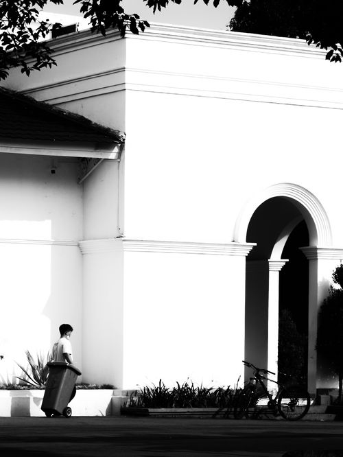 Free Black and white image of a person with a trash bin near an arched building. Stock Photo