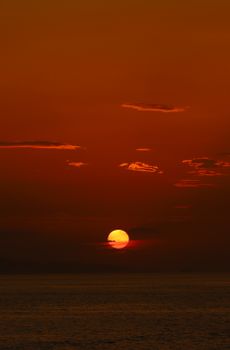 Setting sun over calm sea under orange-red dusk sky with wispy clouds