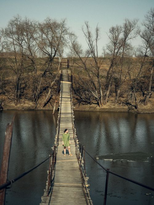 Free A solitary figure walks on a rustic wooden footbridge over a calm river. Stock Photo