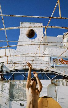 A shirtless man throws a basketball in midair against a clear blue sky.