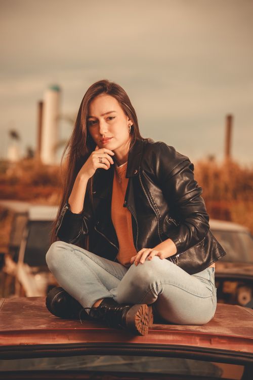 Free A woman wearing a black leather jacket sits pensively on a car hood, exuding a bold and introspective vibe. Stock Photo