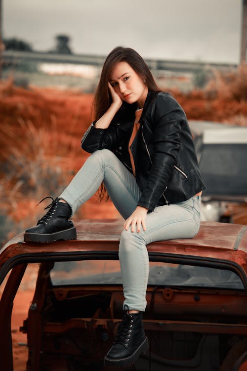 Woman in Black Leather Jacket ad Denim Pants Sitting on Top of a Junked Car