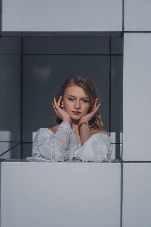 Free A young woman poses gracefully in a white dress, framed by architectural lines, creating a striking portrait. Stock Photo