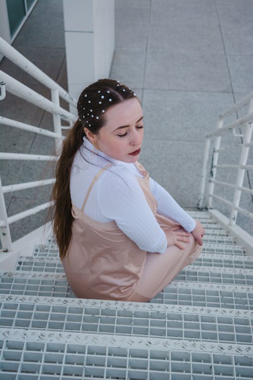 Woman Closing Her Eyes in White Long Sleeve Shirt Sitting on an Outdoor Steel Staircase
