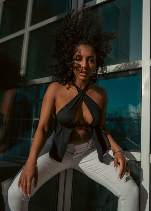Beautiful curly, dark hair, African American woman modeling in front of reflected windows, wearing a black criss crossed top with white pants.
