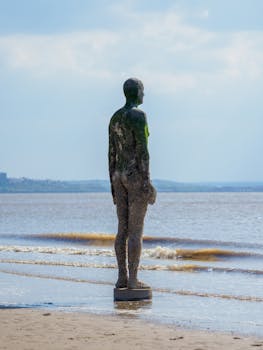 A solitary sculpture on Crosby Beach against a serene ocean backdrop under a blue sky.