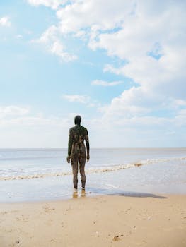 A statue stands on Crosby Beach, England, facing the serene sea under a clear sky.