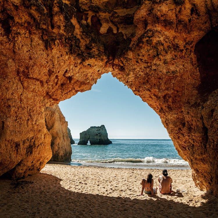 Young Couple Sitting On Beach Under Big Rocky Arch