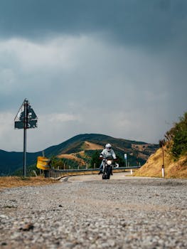 A motorcyclist travels along a curvy mountain road under a dramatic sky.