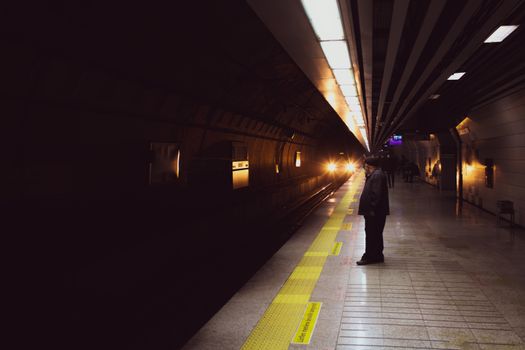 A person waits on a subway platform in Istanbul as a train approaches in the dimly lit station.
