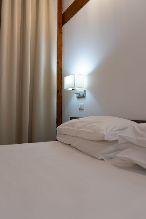 White Bed Linen and Pillows Near a Wall Lamp