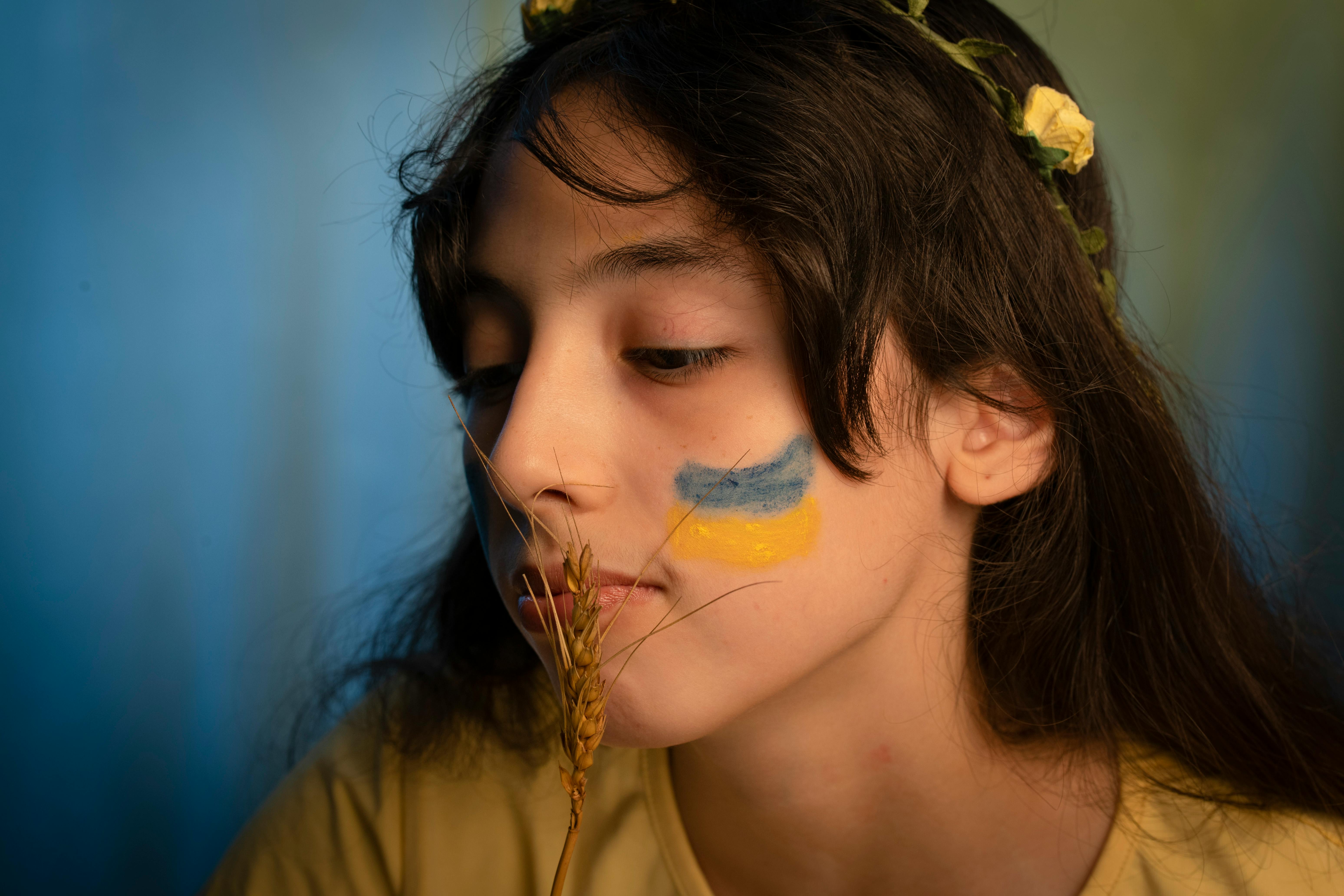 Young girl with floral wreath, wheat, and blue-yellow face paint in a peaceful setting.