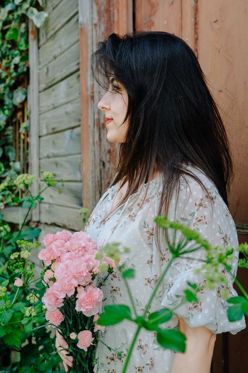 Free Side view of a woman holding pink flowers near wooden structure amidst greenery. Stock Photo