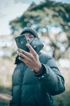 A young man wearing a jacket holds two smartphones outdoors, showcasing modern communication.