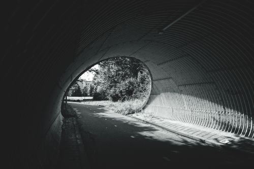 Free A black and white photo of a tunnel leading to a sunlit path with trees and shadows. Stock Photo