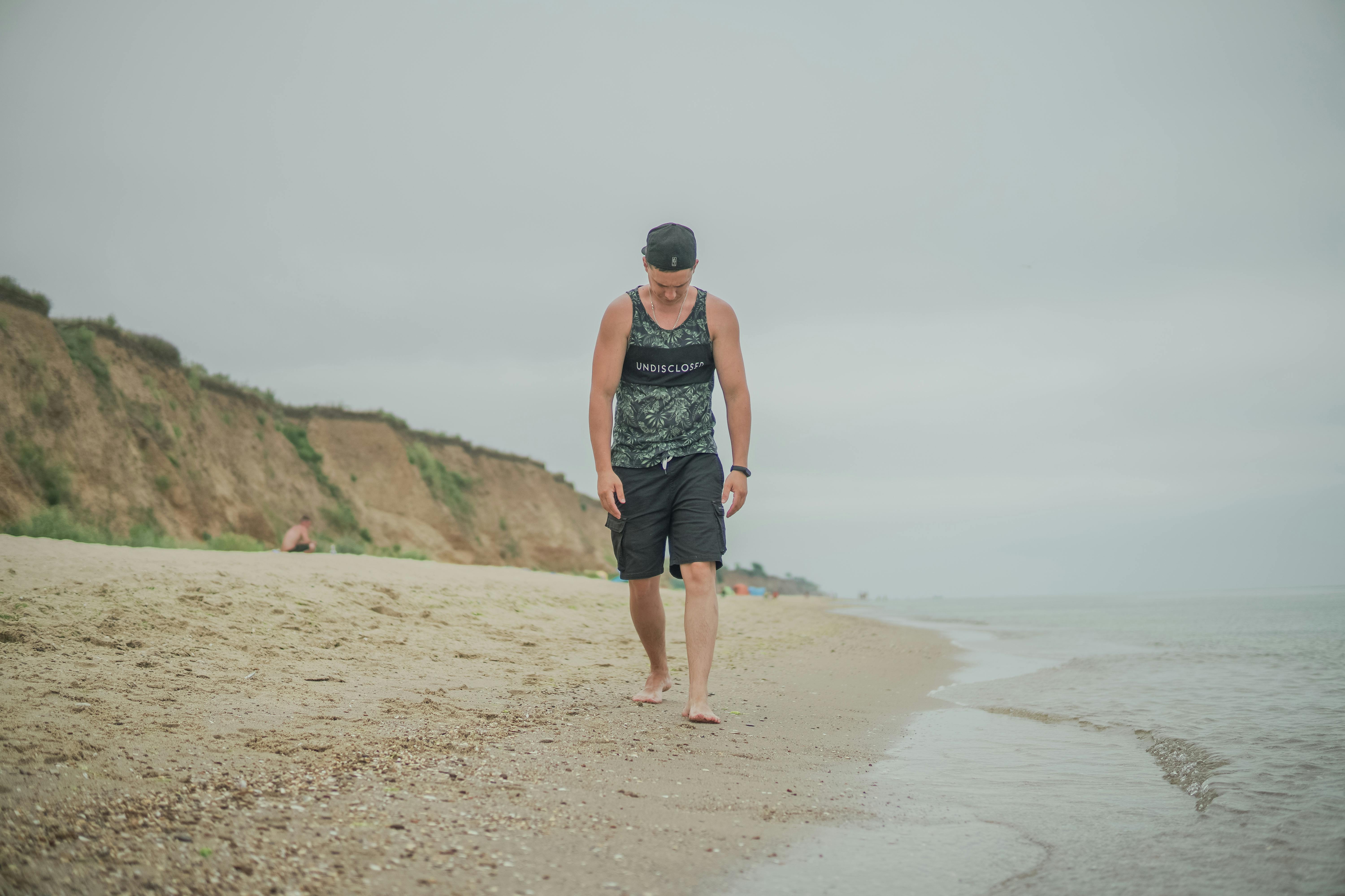 Casual man in tank top and cap walking barefoot on Odesa beach, Ukraine.