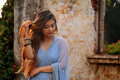 A woman in a blue saree poses gracefully outdoors by a rustic wall.