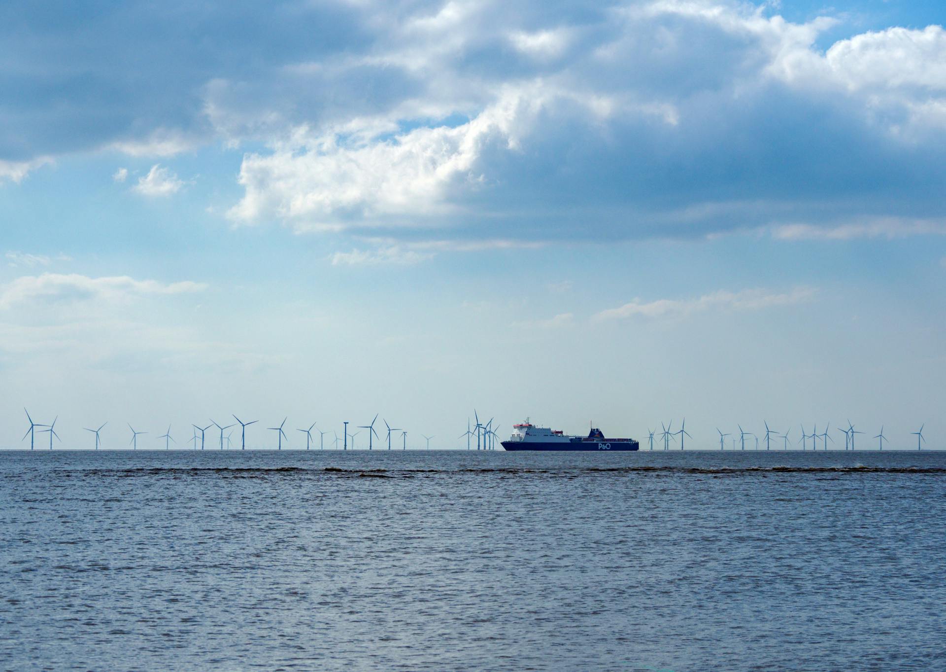 A ferry passing by offshore wind turbines in the sea under a cloudy sky in England.