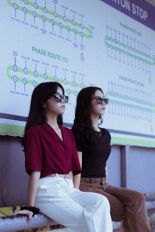 Women in Red and Black Shirt Wearing Sunglasses Sitting on the Street with Their Legs Crossed