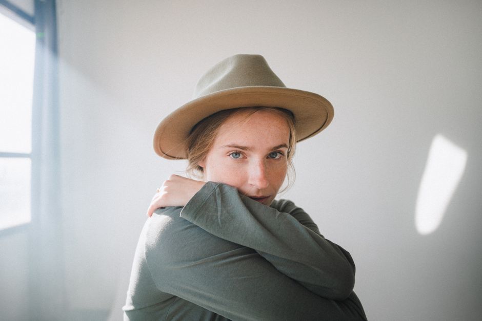 Soft portrait of a woman hugging herself in a sunlit room, wearing a stylish hat.