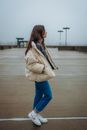 Woman in White Coat and Blue Denim Jeans Walking on Gray Concrete Pavement