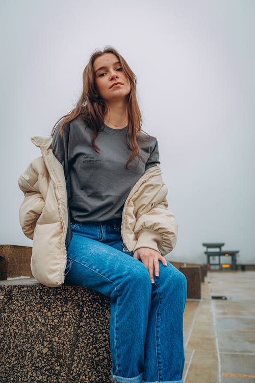 A Woman in Denim Jeans Looking Down while Sitting on a Concrete Surface