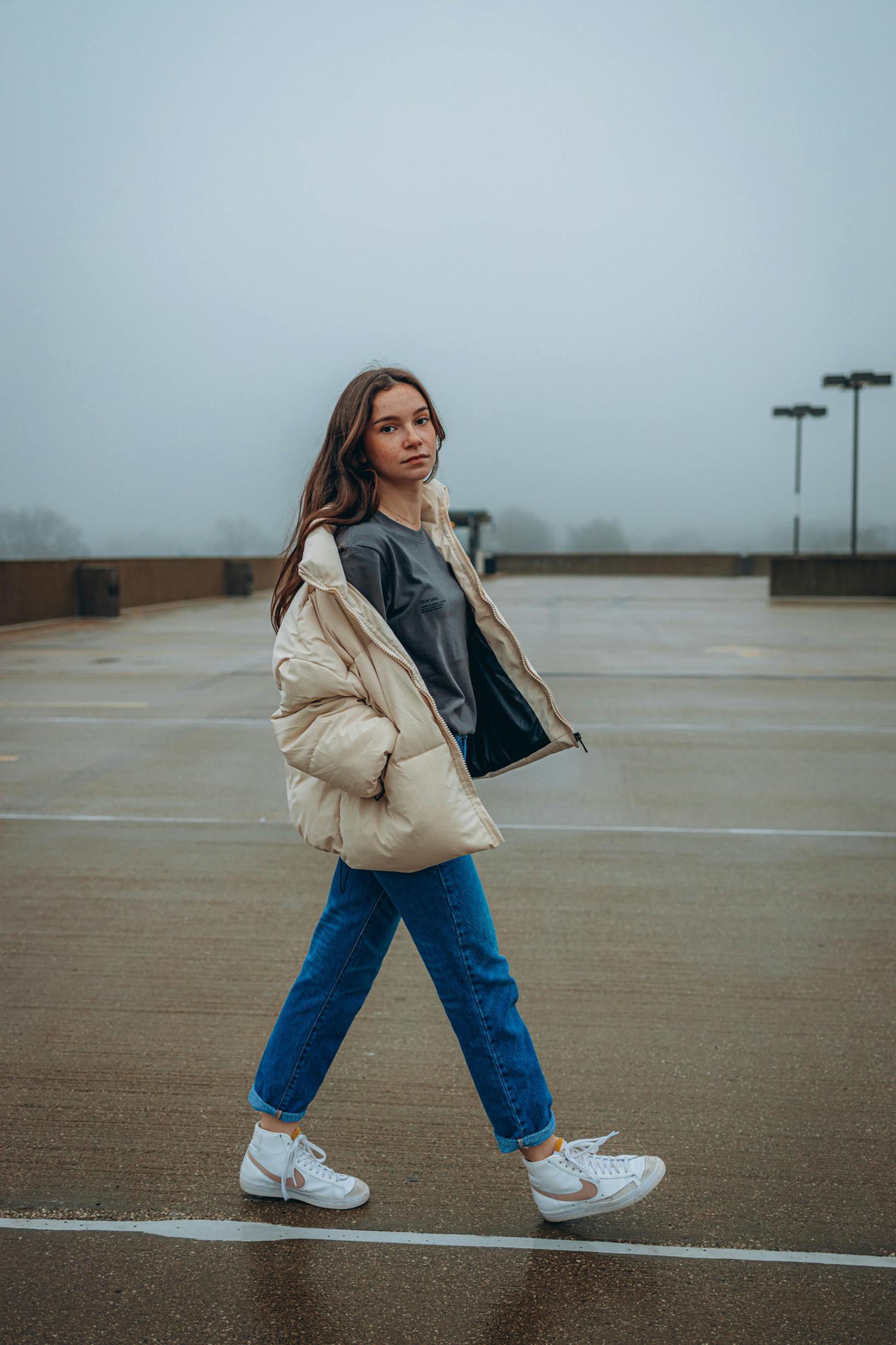 A young woman in casual attire confidently posing on a foggy rooftop parking lot.